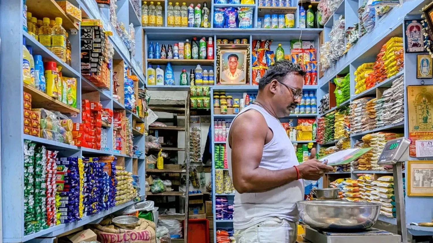 A kirana shopkeeper weighing goods on the shop floor, surrounded by densely stocked shelves